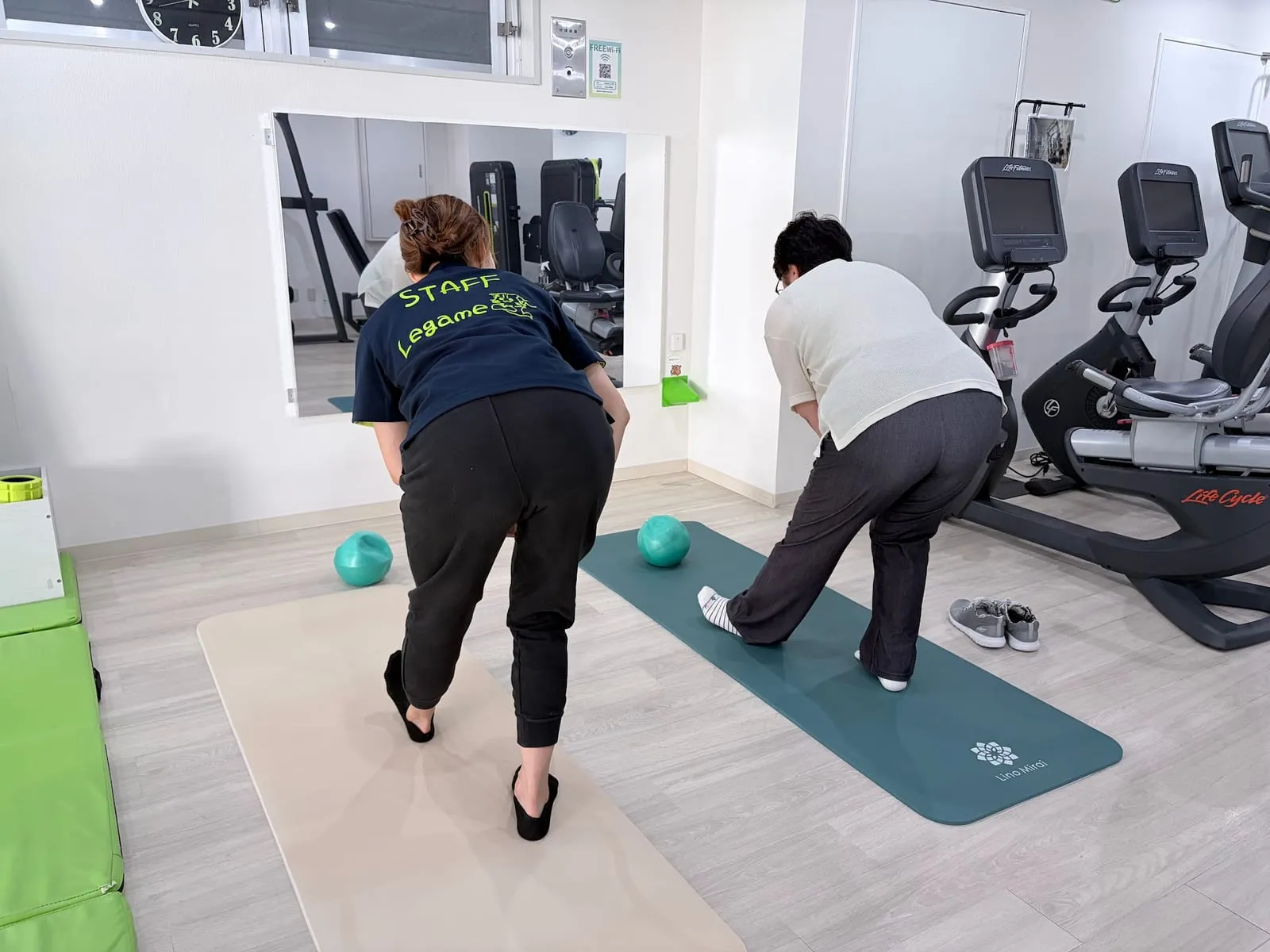 スタッフと会員が並んで前屈の動きをしている（後ろ姿）